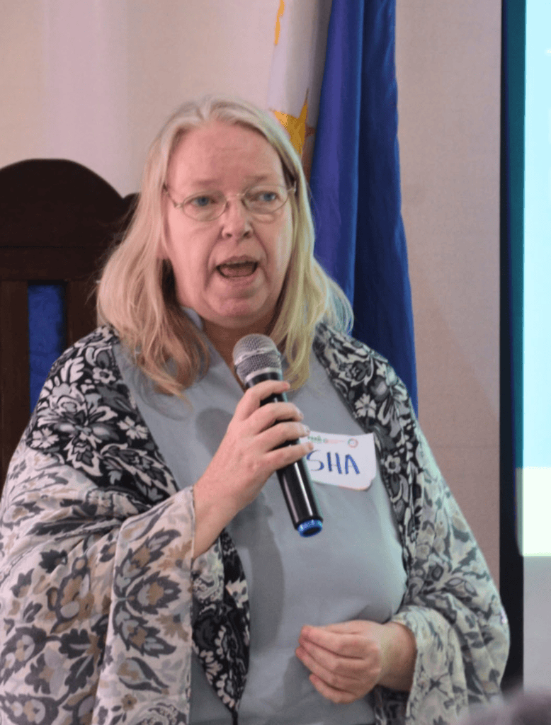 Photo of Dr. Sharon Taylor with a gray shirt and a pretty scarf speaking on an event with the Philippine flag behind her.